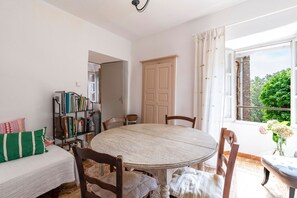 Ein Zimmer mit rundem Tisch, Stühlen, einem Bücherregal, einem Bett und einem Fenster mit Vorhängen.