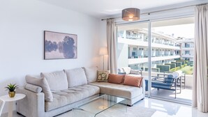 Een moderne woonkamer met een bank, een glazen salontafel, een schilderij en uitzicht op een wooncomplex.