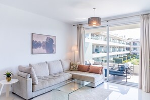 A modern living room with a sofa, a glass coffee table, a painting, and a view of a residential building. - Cubo's Bel-Air Estepona Apartment / communal pool / parking included / air conditioning / free Wi-Fi (Estepona)