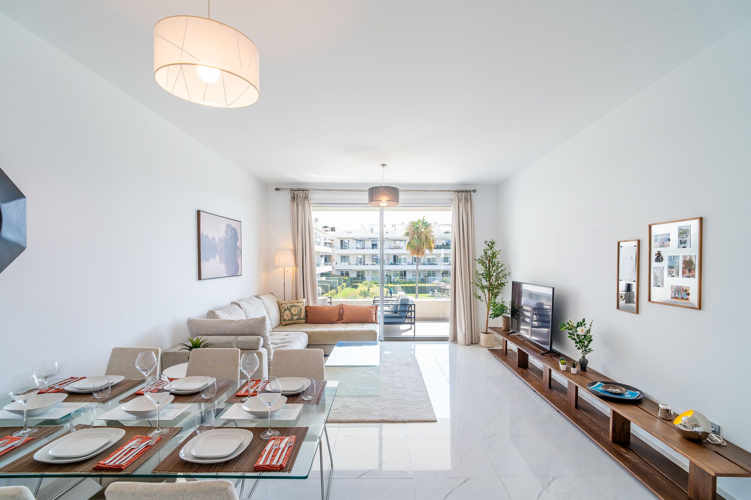A modern living room with a dining area, a sofa, a TV, and a balcony with a view of buildings.
