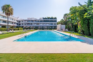 Une piscine rectangulaire à l’eau d’un bleu limpide, entourée d’une terrasse dallée avec des chaises longues et des parasols. Au fond, on aperçoit un immeuble à plusieurs étages doté de balcons et un jardin luxuriant.