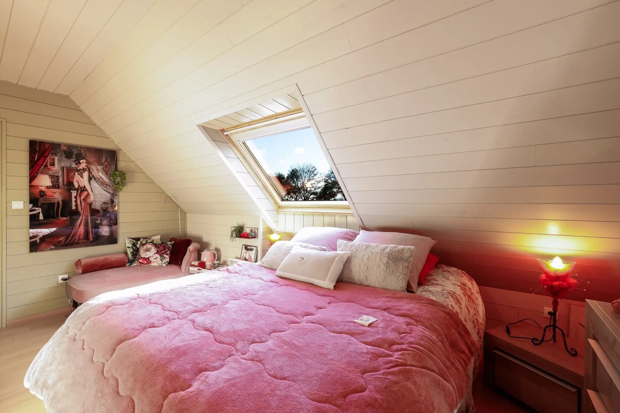 Quarto com teto inclinado, claraboia, cama grande com roupa de cama rosa, mesa de cabeceira com abajur e um quadro na parede.