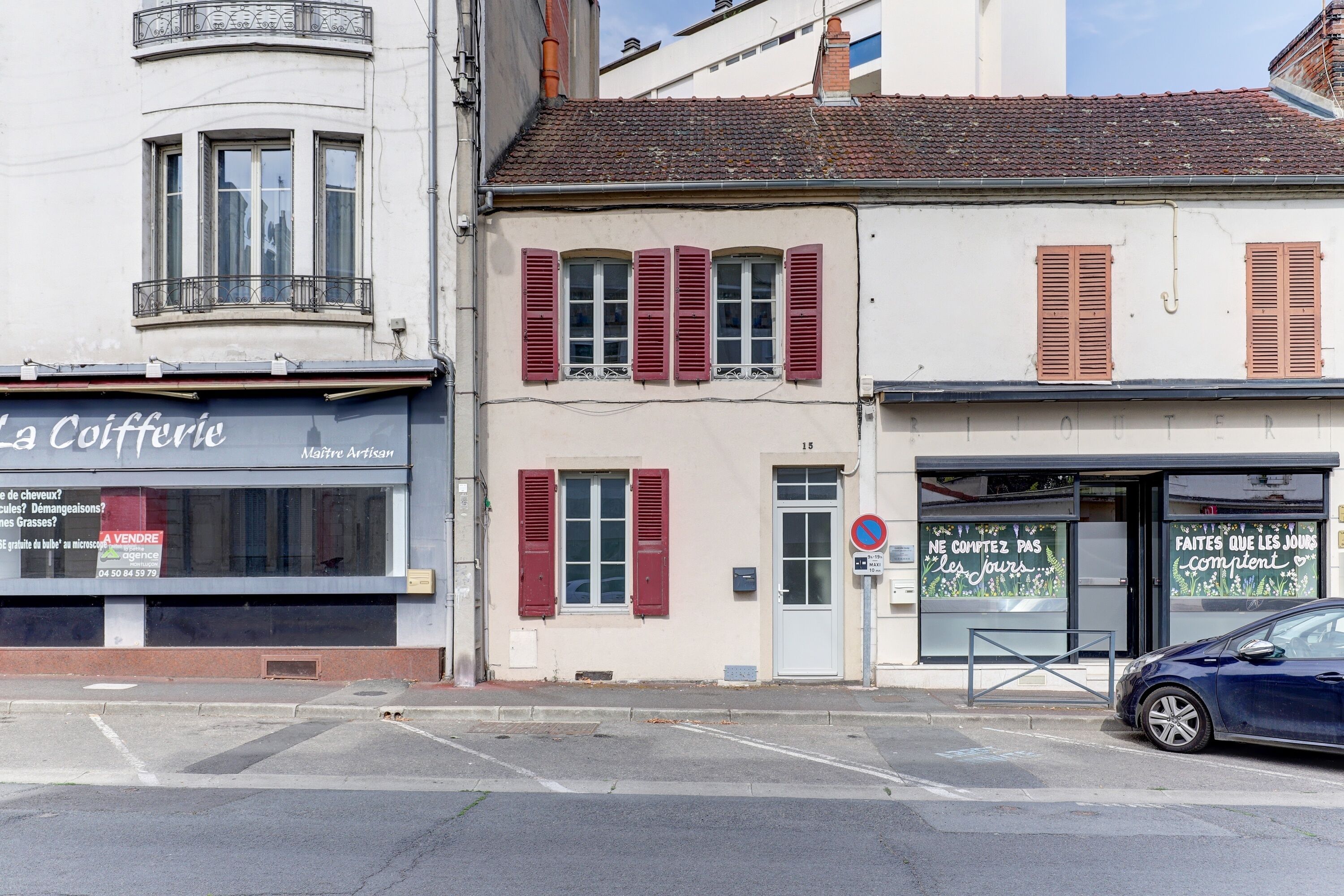 A street view with a café named 'La Cofferie' on the left, a building with red shutters in the middle, and a blue car parked on the right.