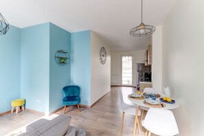 A modern living room with a blue accent wall, a dining area with a white table and chairs, and a kitchen in the background. - Le Cocon Bleu - Bel appt pour 6 en centre-ville (Montlucon)