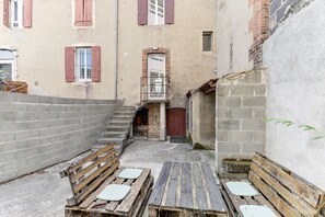 Ein schmaler Gang mit einer Holzbank und einem kleinen Tisch, einer Backsteinmauer und einem Gebäude mit roten Fensterläden.