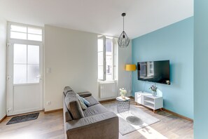 A modern living room with a sofa, a TV mounted on the wall, and a small side table with a lamp and flowers. - Le Cocon Bleu - Bel appt pour 6 en centre-ville (Montlucon)