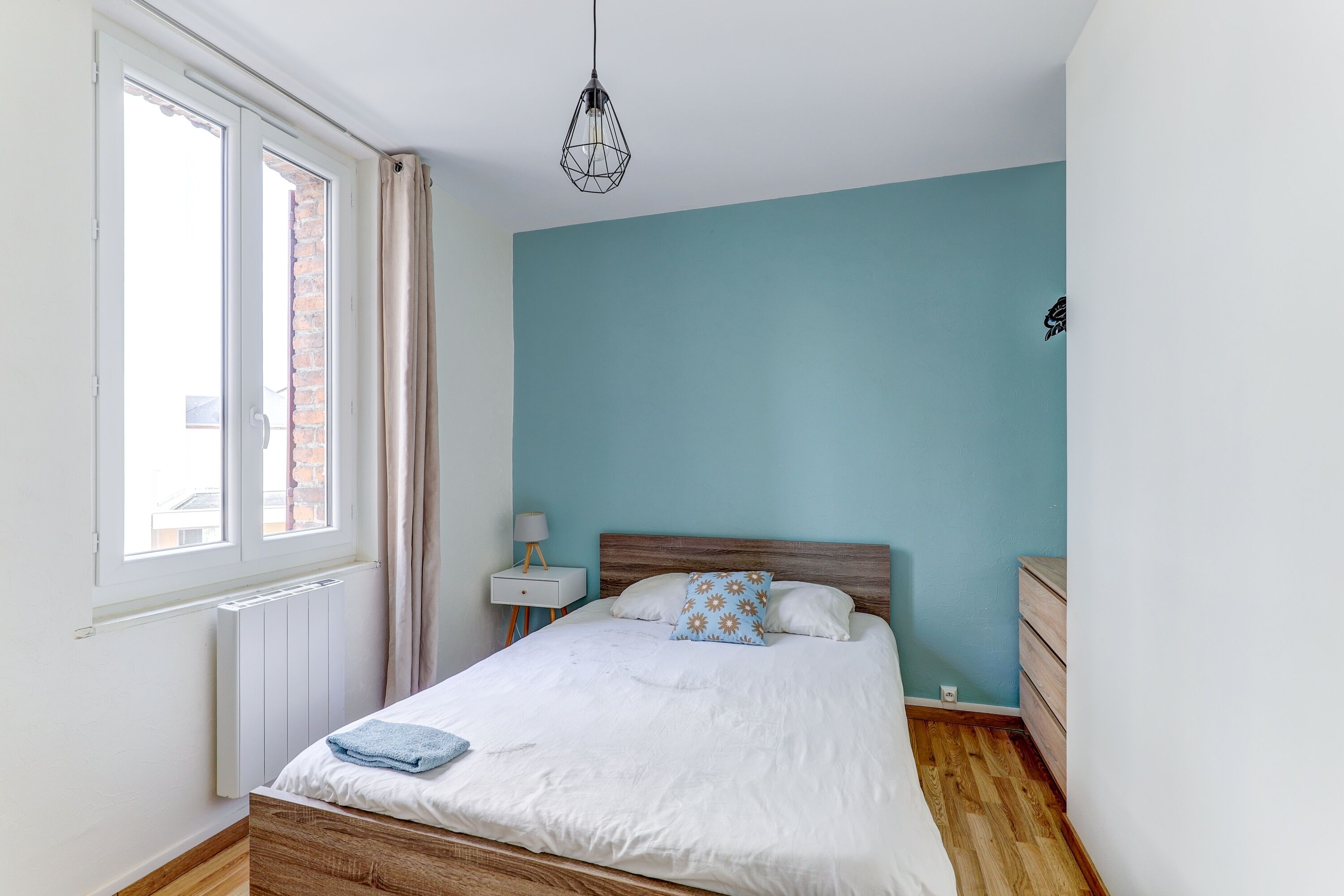 Une chambre à coucher avec un lit, une table de chevet, une lampe, une fenêtre et un luminaire suspendu.