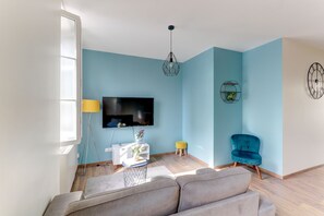 Uma sala de estar com sofá, televisão, parede azul, luminária pendente, uma mesinha com um vaso e uma cadeira.