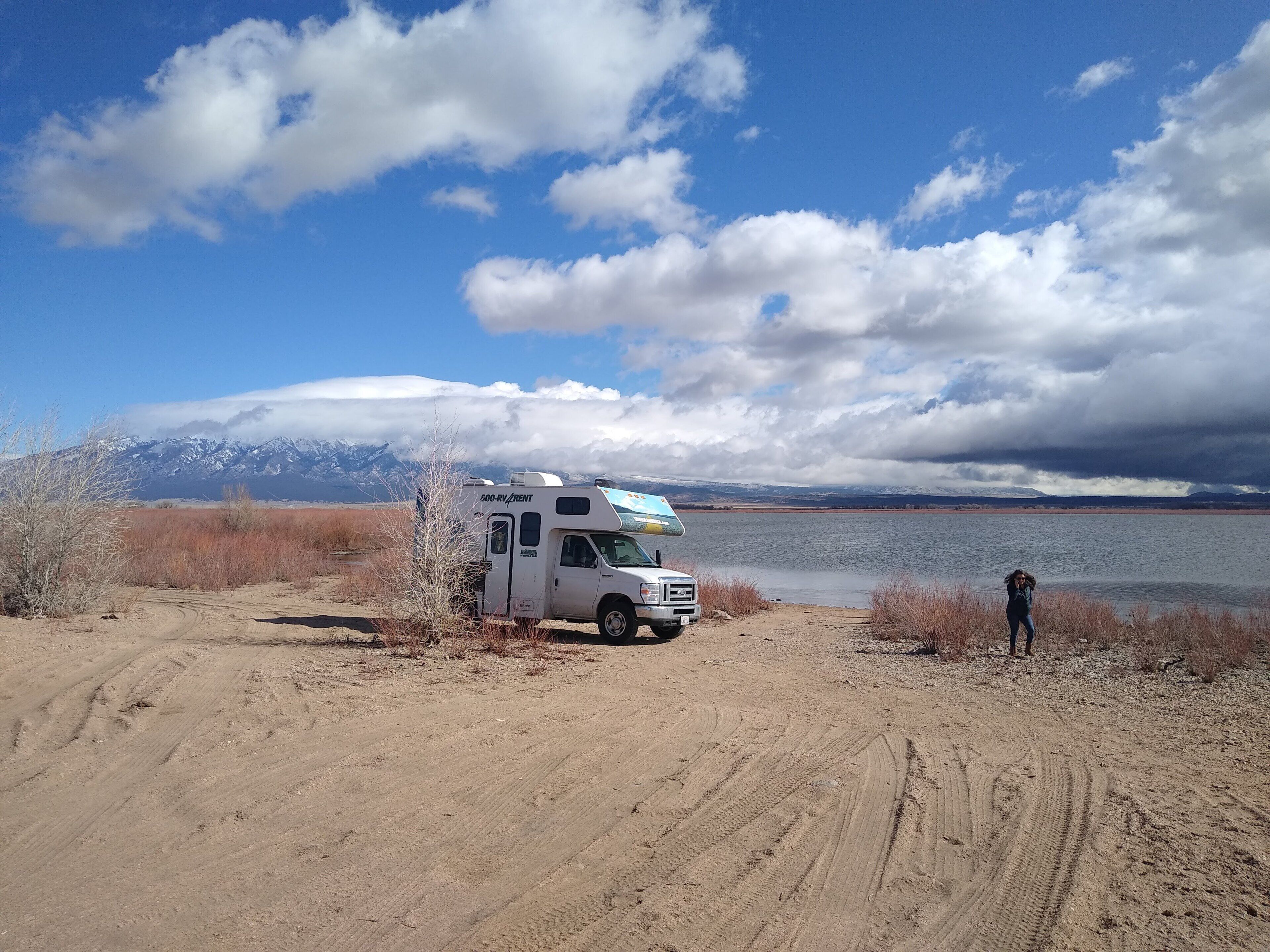 A white RV parked on a dirt path with a lake and mountains in the background.