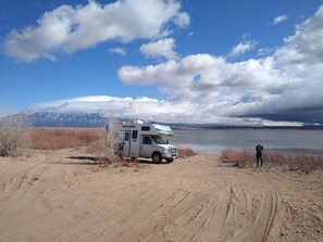 A white RV parked on a dirt path with a lake and mountains in the background.