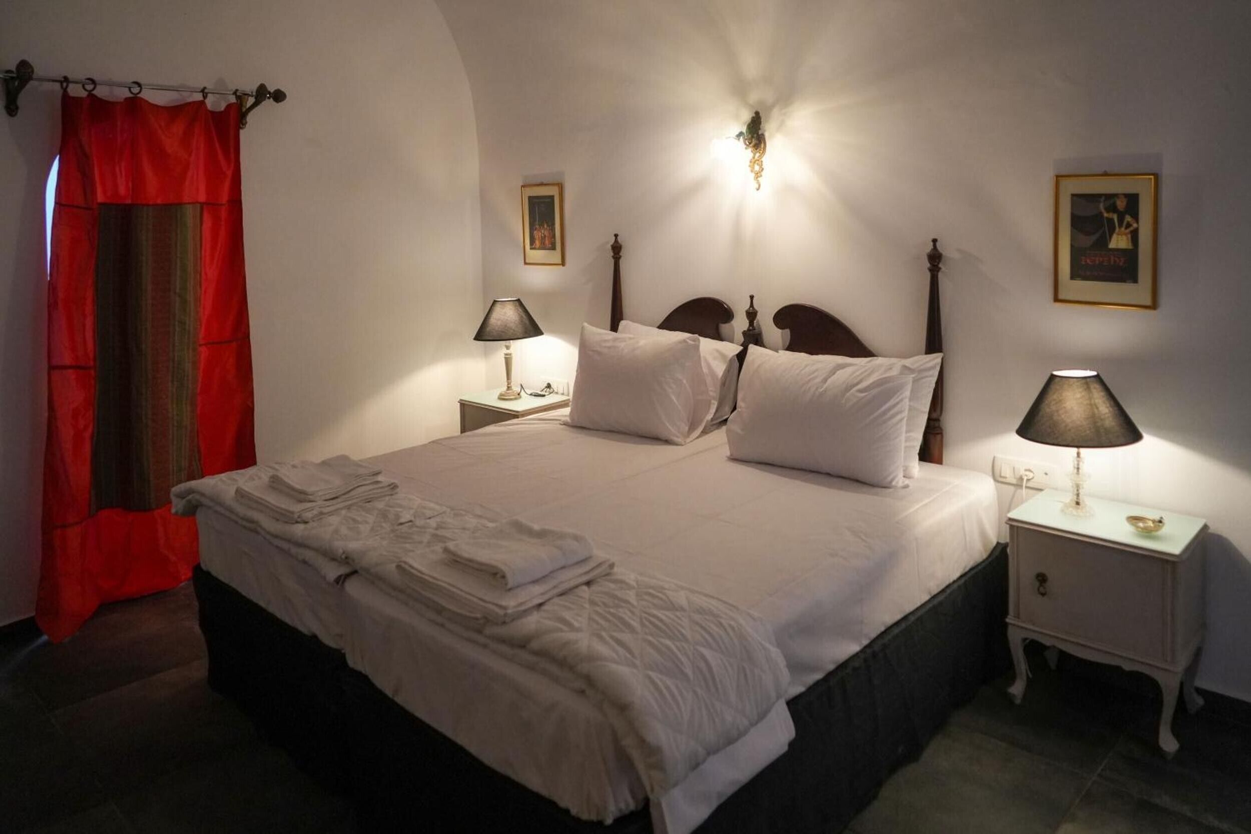Ein ordentlich bezogenes Bett mit Kissen und einem gefalteten Handtuch, eine Nachttischlampe, ein gerahmtes Bild an der Wand und ein roter Vorhang.