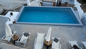 A rectangular pool with a clear blue water surface, surrounded by white lounge chairs and umbrellas, set against a concrete deck with a sandy area in the background. - Apartment 'Two-Bedroom' with Wi-Fi and Air Conditioning (Emporio)