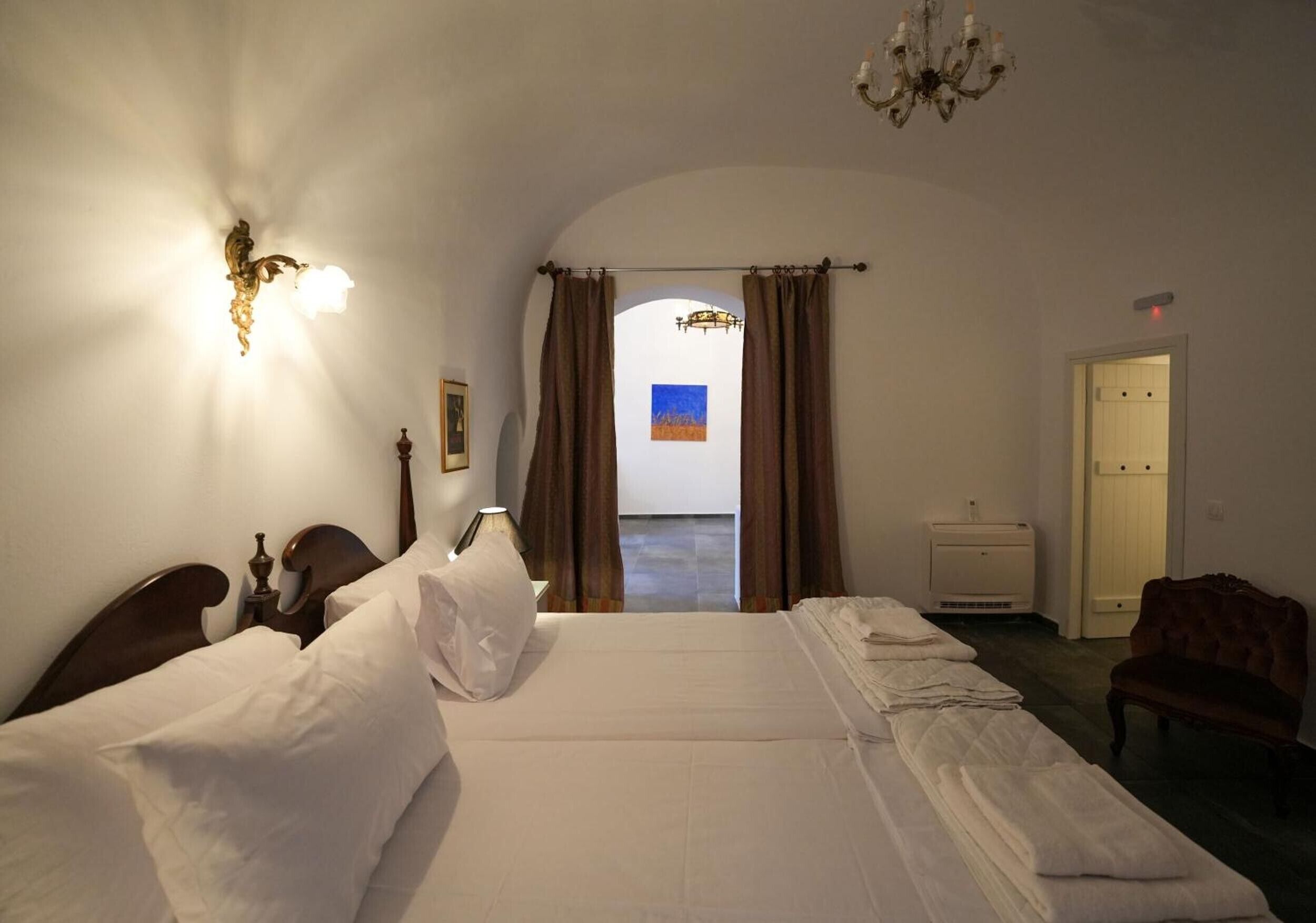 A hotel room with a large bed, a desk, a chair, a chandelier, and a view through a doorway.