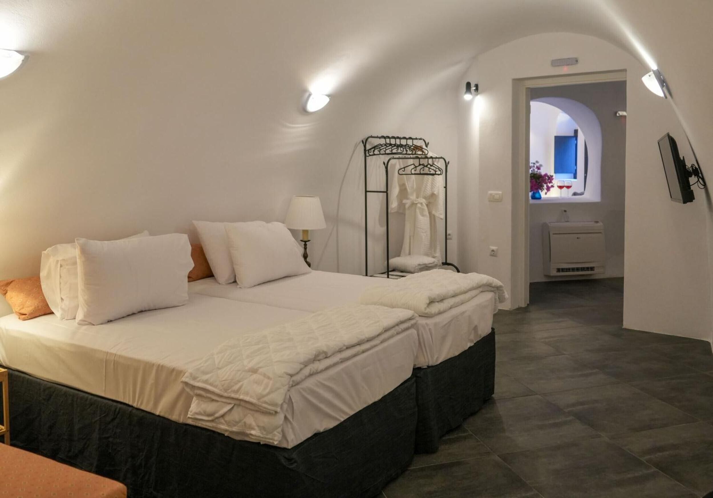 Ein Schlafzimmer mit zwei Betten, einem Nachttisch, einer Lampe, einem Kleiderständer und Blick auf ein Badezimmer durch einen Rundbogen.