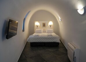 Una estança petita i allargassada, amb volta d'arc, un llit al centre, dues làmpades penjades a la paret, un quadre emmarcat i un televisor penjat a la paret.
