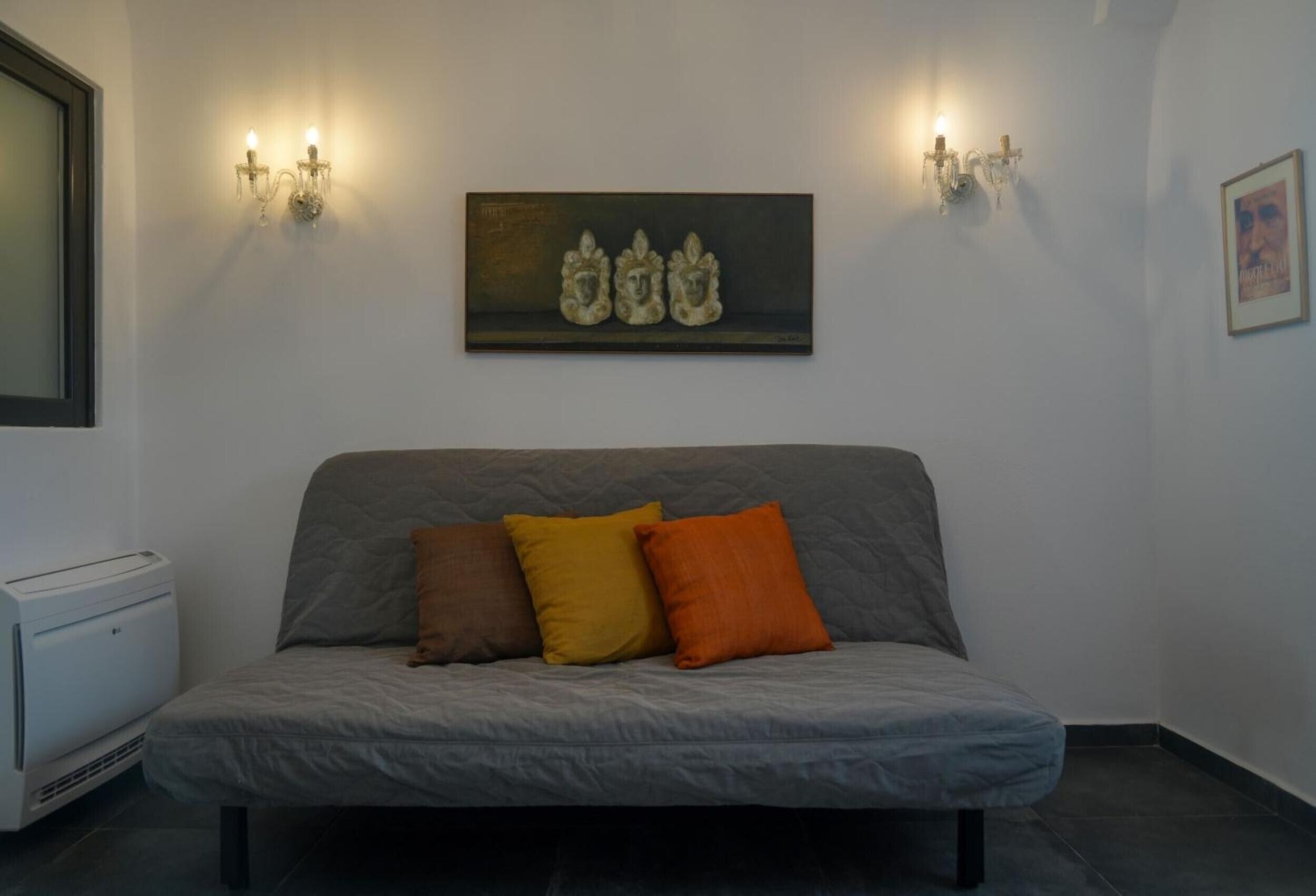 A room with a grey sofa, three cushions, a painting above the sofa, two wall-mounted lights, a framed picture on the wall, and a white device on the floor.