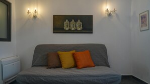 Une pièce avec un canapé gris, trois coussins, un tableau au-dessus du canapé, deux lampes fixées au mur, une photo encadrée accrochée au mur et un appareil blanc posé sur le sol.