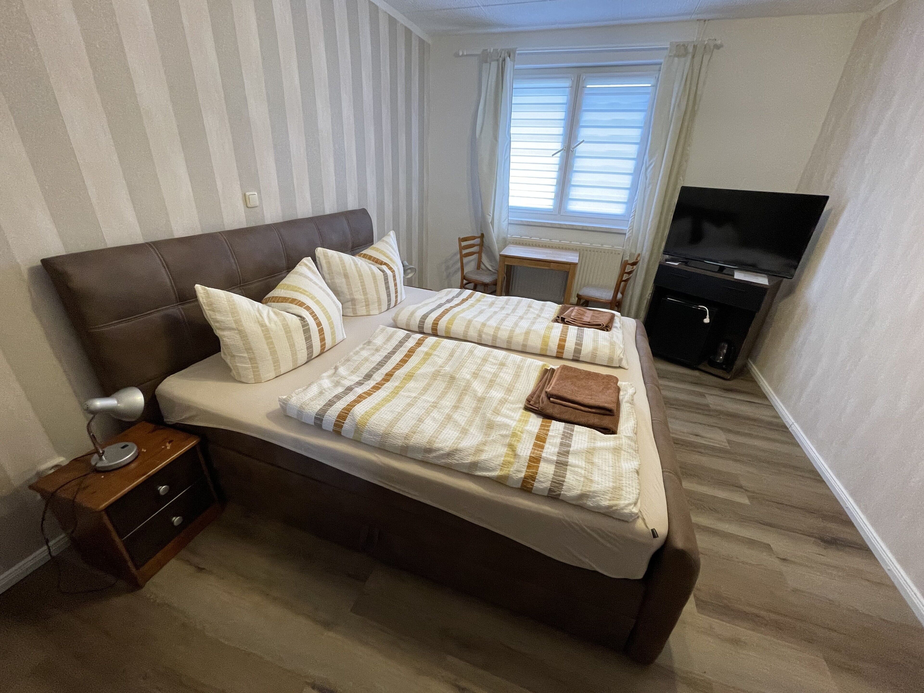 Ein Doppelbett mit gestreifter Bettwäsche, ein Nachttisch mit Lampe, ein Fernseher und ein kleiner Schreibtisch mit Stuhl.