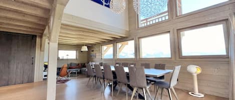 A spacious dining area with a long table and chairs, large windows, and a high ceiling with exposed beams.