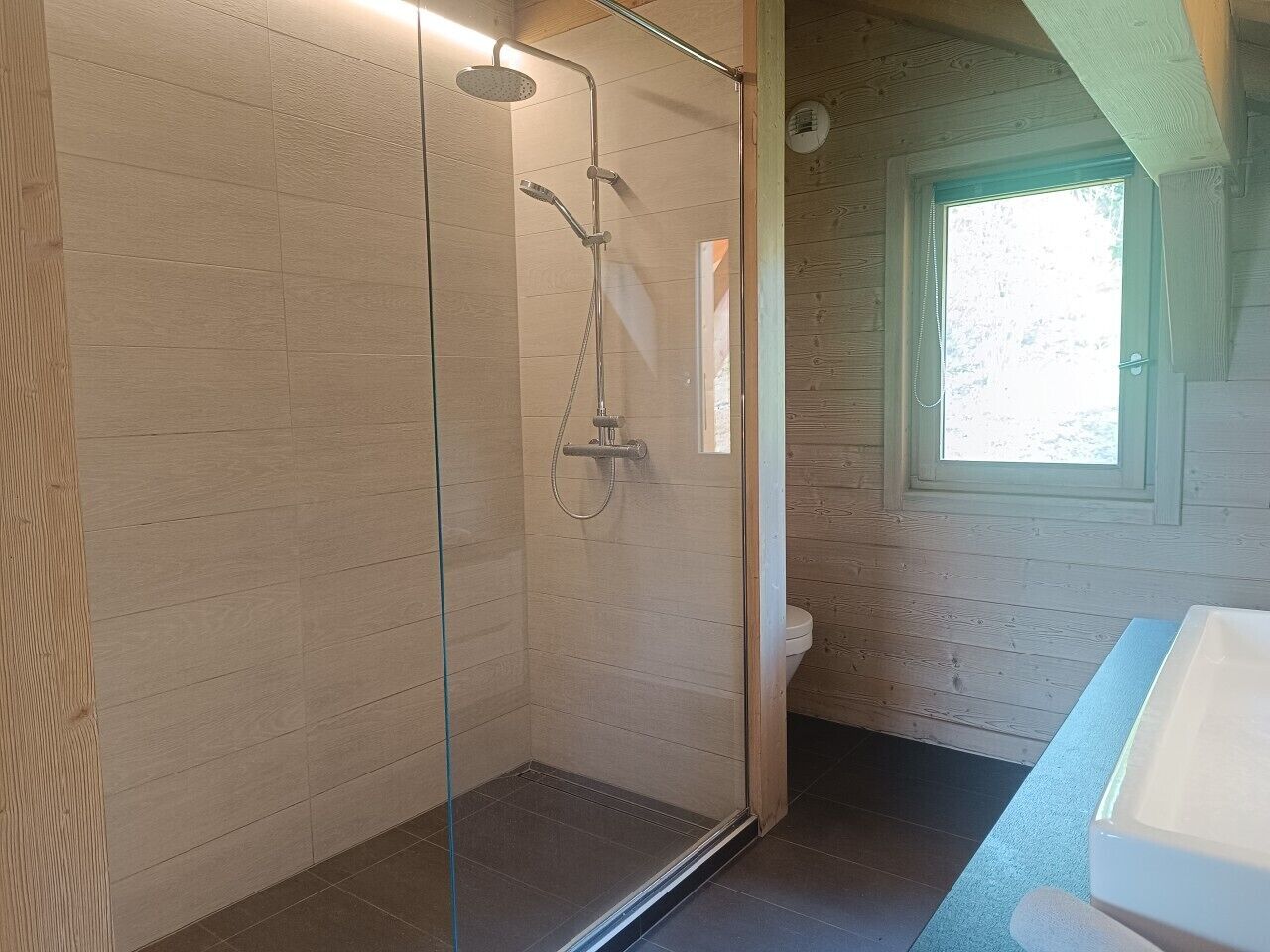 A modern bathroom with a glass-enclosed shower, a wooden wall, and a window.