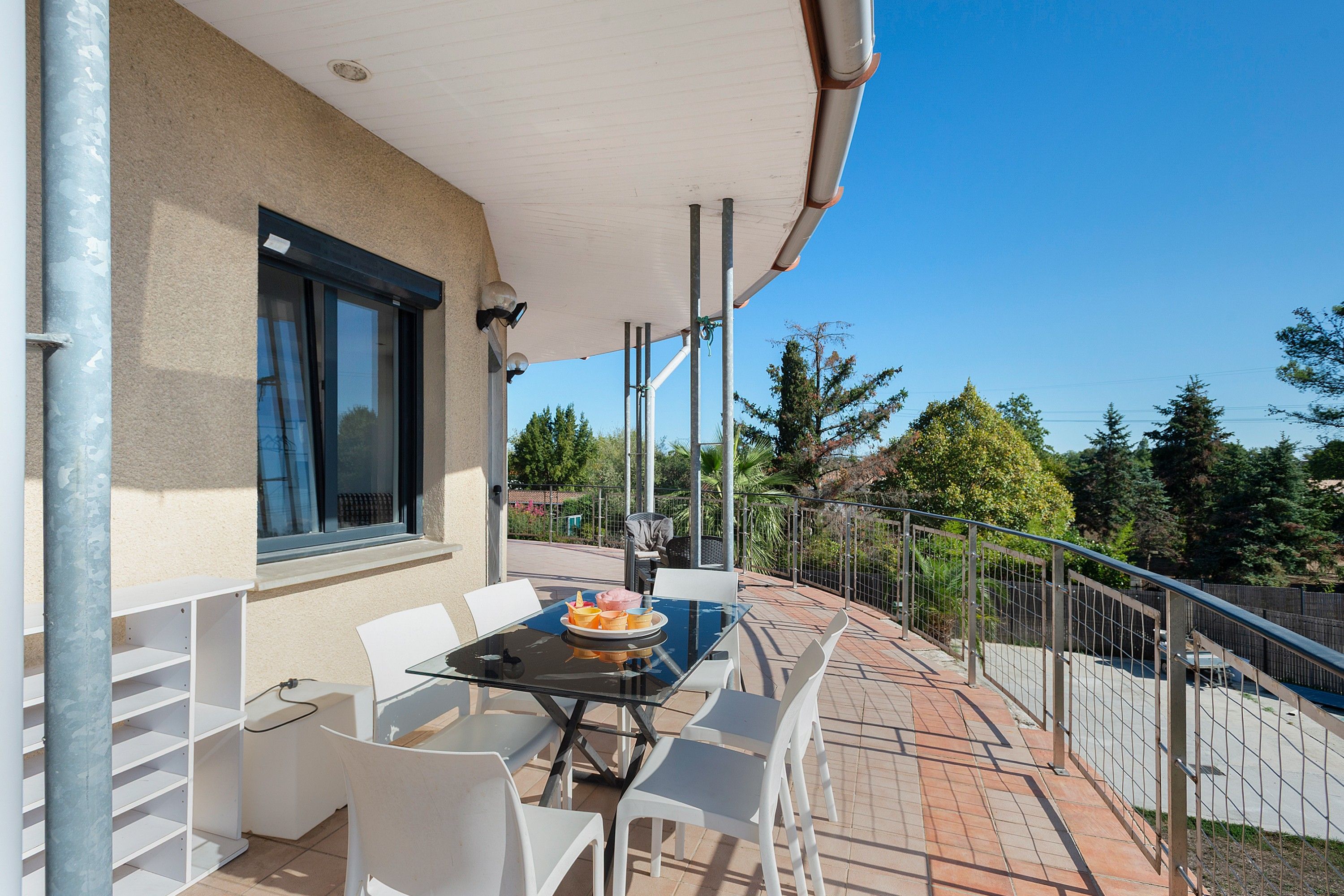 An outdoor patio with a glass table and white chairs, a window with blinds, and a view of trees and a fence.