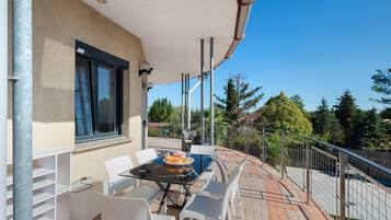 An outdoor patio with a glass table and white chairs, a window with blinds, and a view of trees and a fence.