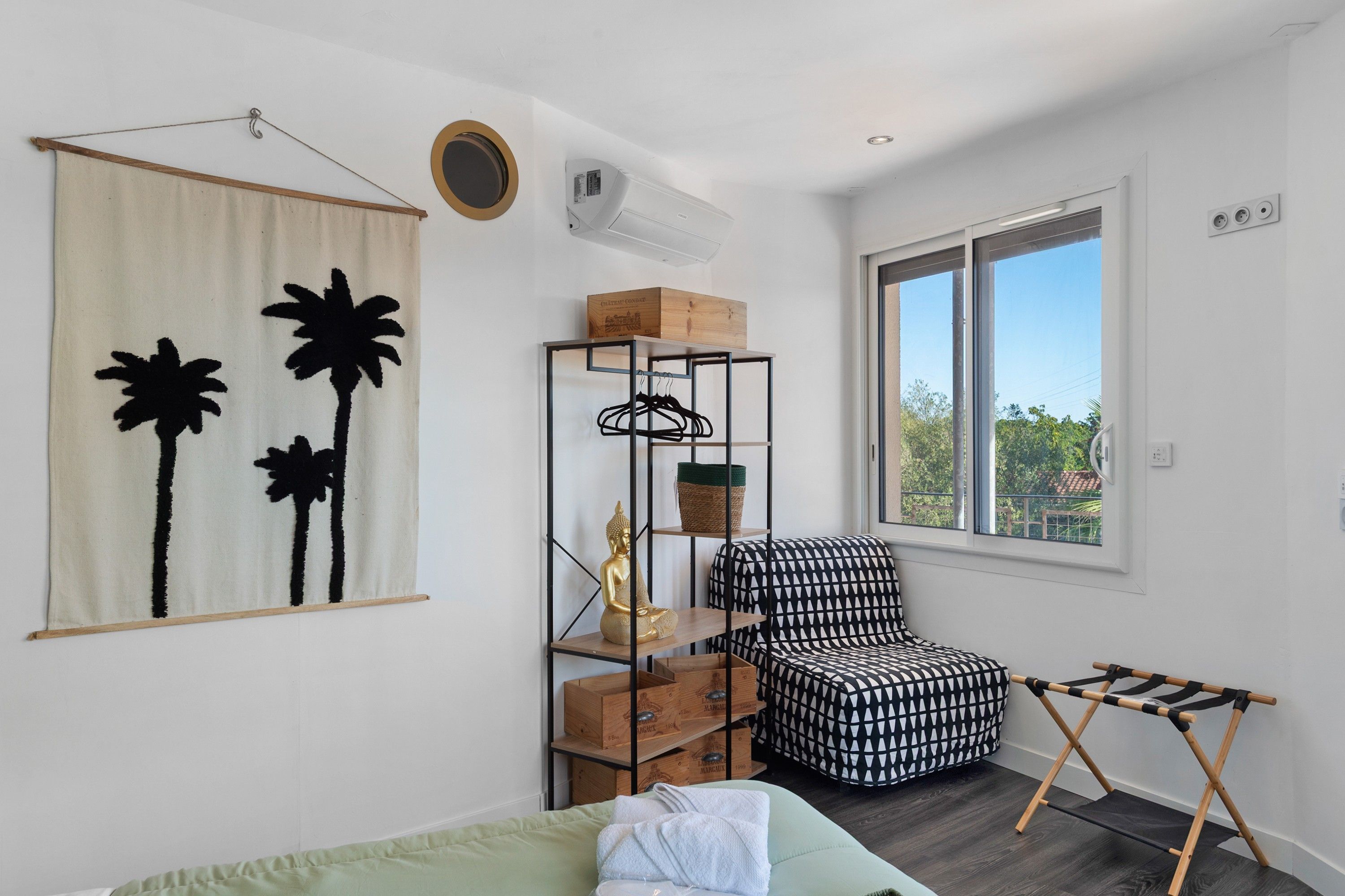 Une pièce comprenant un lit, une chaise, une fenêtre, une étagère fixée au mur avec des boîtes et une tapisserie suspendue.