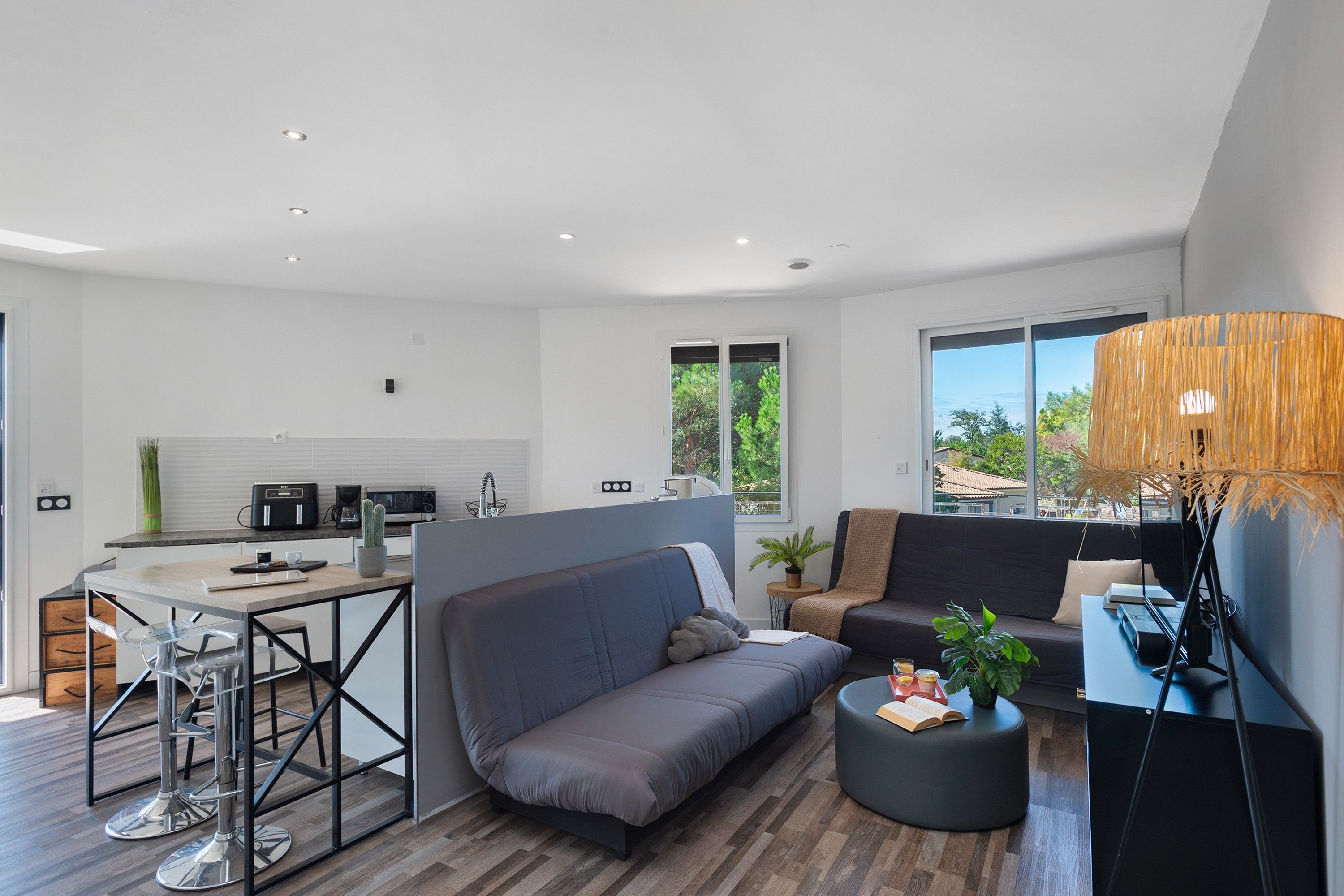 A modern living space with a kitchen, a grey sofa, a round ottoman, and a large window with a view.