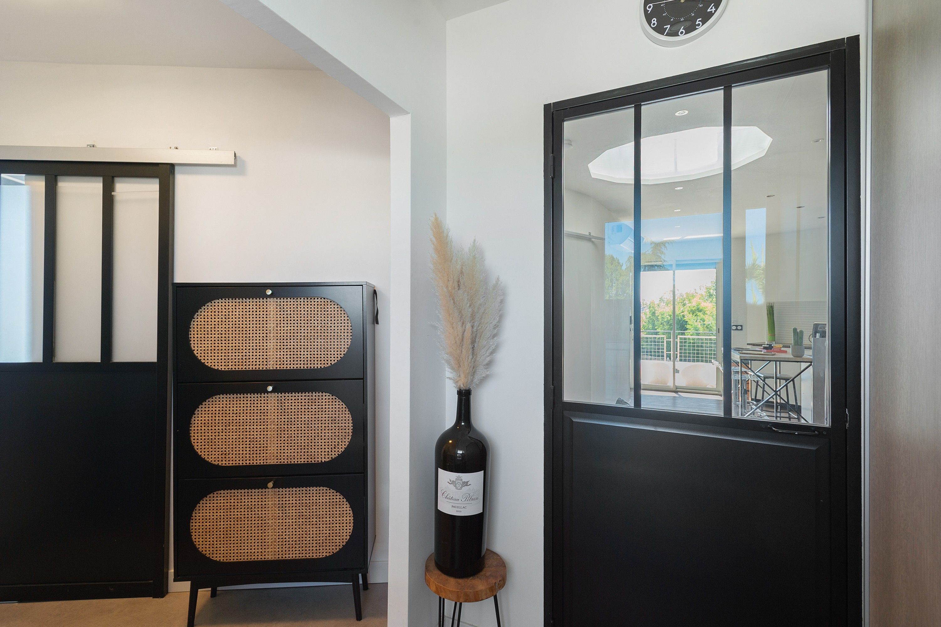A room with a black door, a wall-mounted clock, and a decorative wicker console with a bottle of wine.