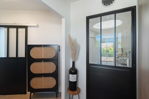 Una habitación con una puerta negra, un reloj en la pared y una consola de mimbre decorativa con una botella de vino.