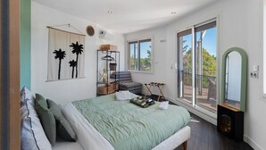 Ein Schlafzimmer mit Bett, einem Sessel, einem kleinen Tisch, einem Spiegel und Blick ins Freie durch eine Schiebetür.