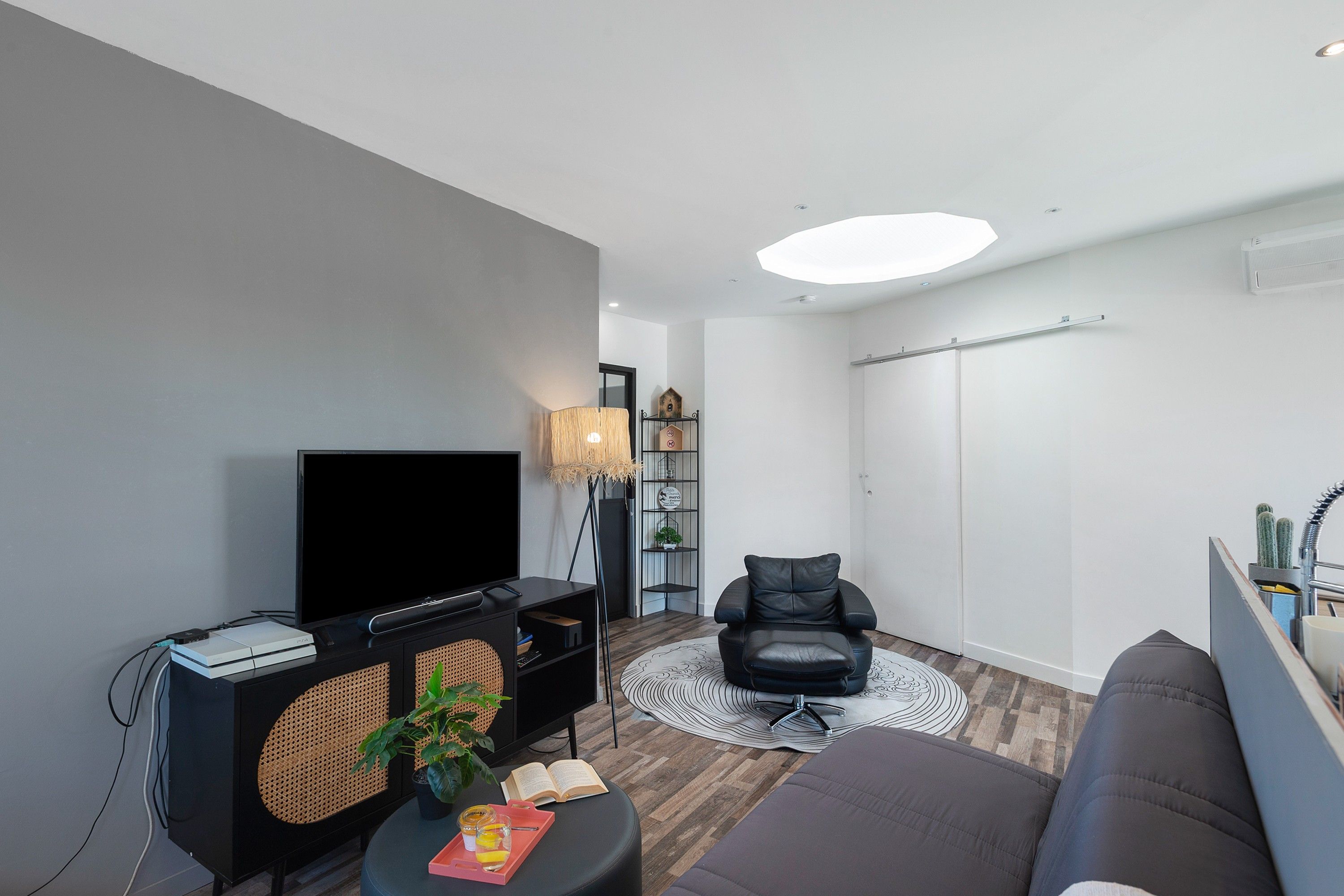 A modern living room with a flat-screen TV, a black leather chair, a sofa, a small table with a plant, and a bookshelf.