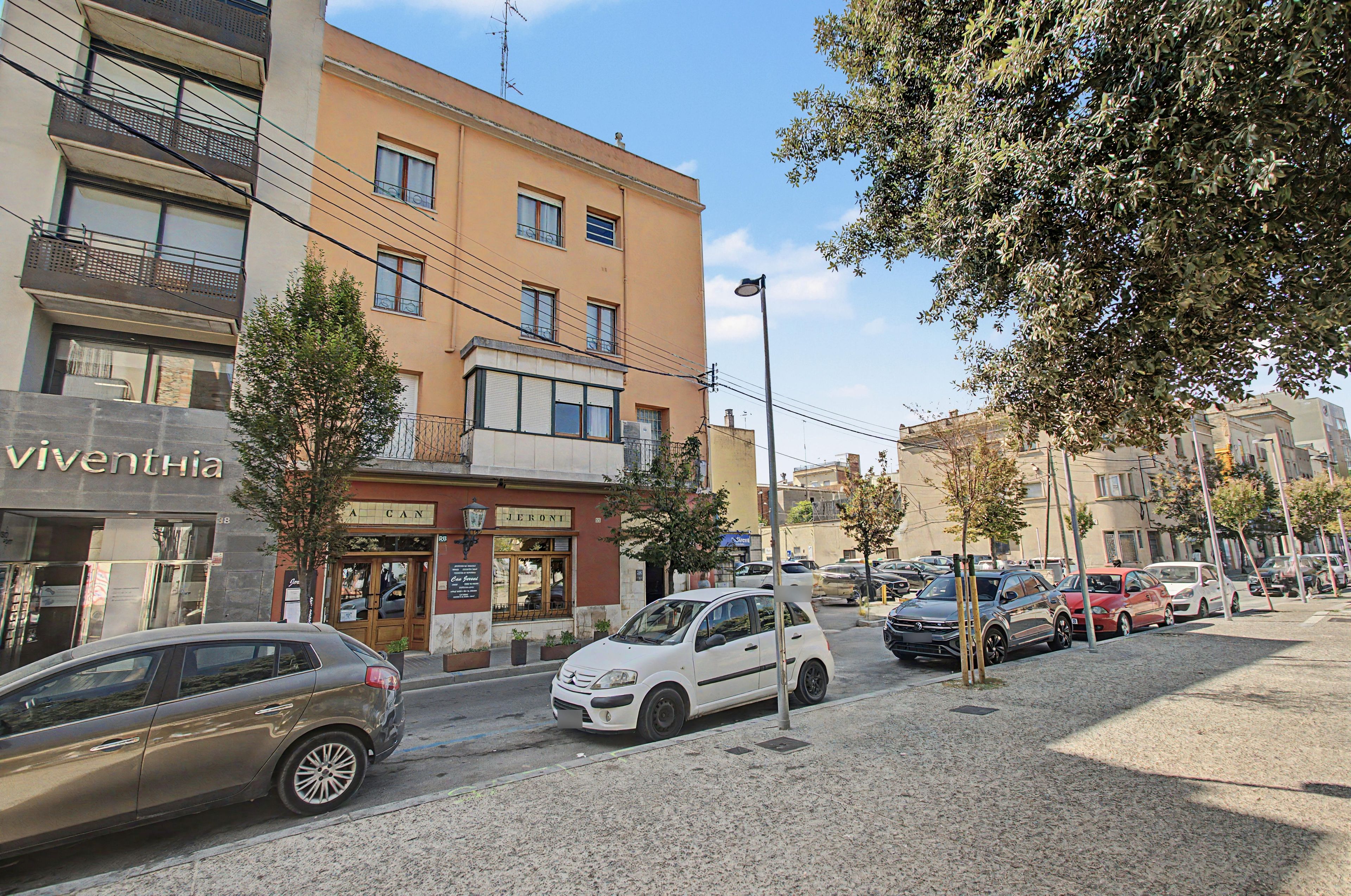 Una calle con coches estacionados, un edificio con una tienda llamada 'A CAN' y 'JERONI', y un árbol en primer plano.