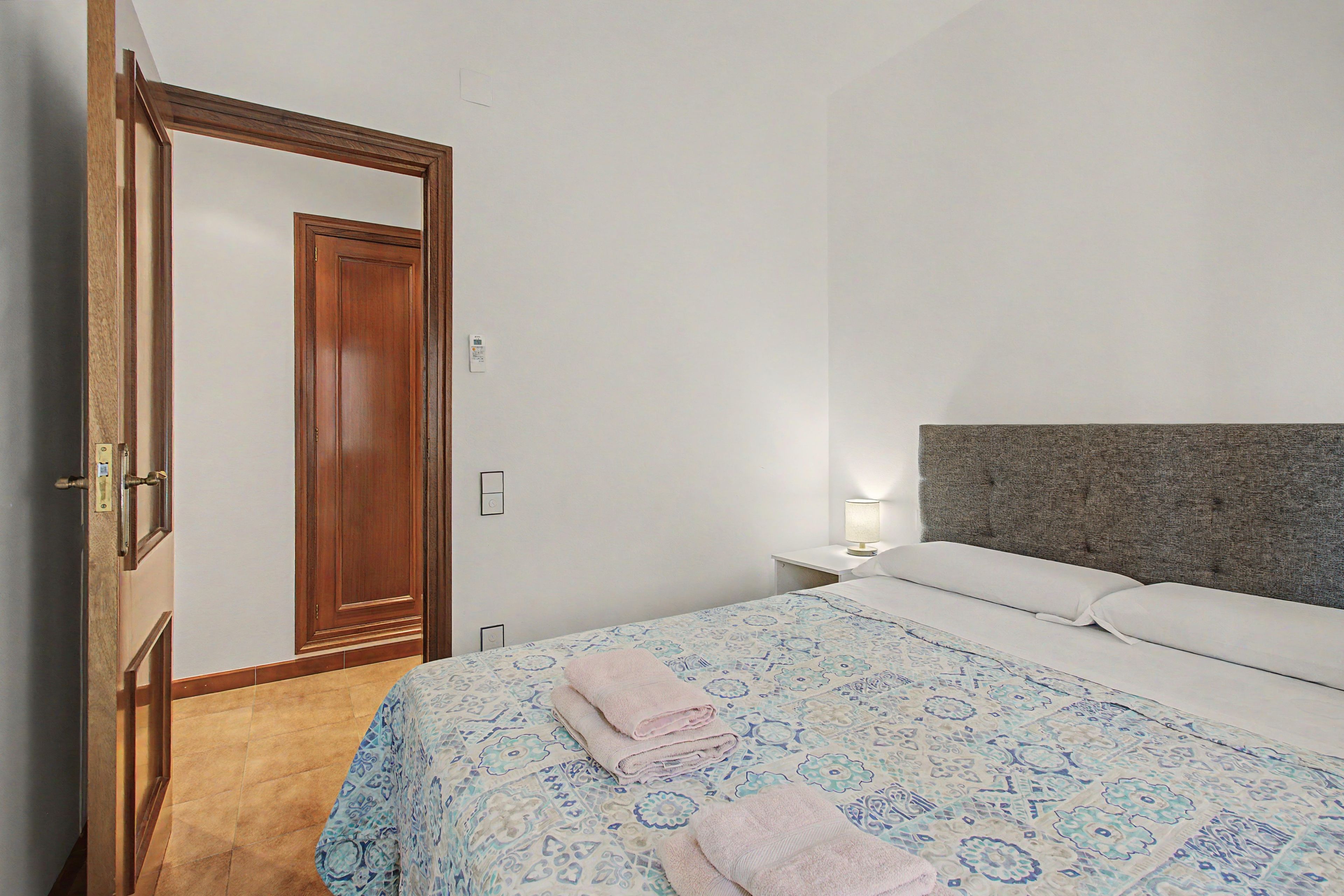 Hotellihuone, jossa on sänky, yöpöytä, lamppu, puuovi ja seinäkytkin.