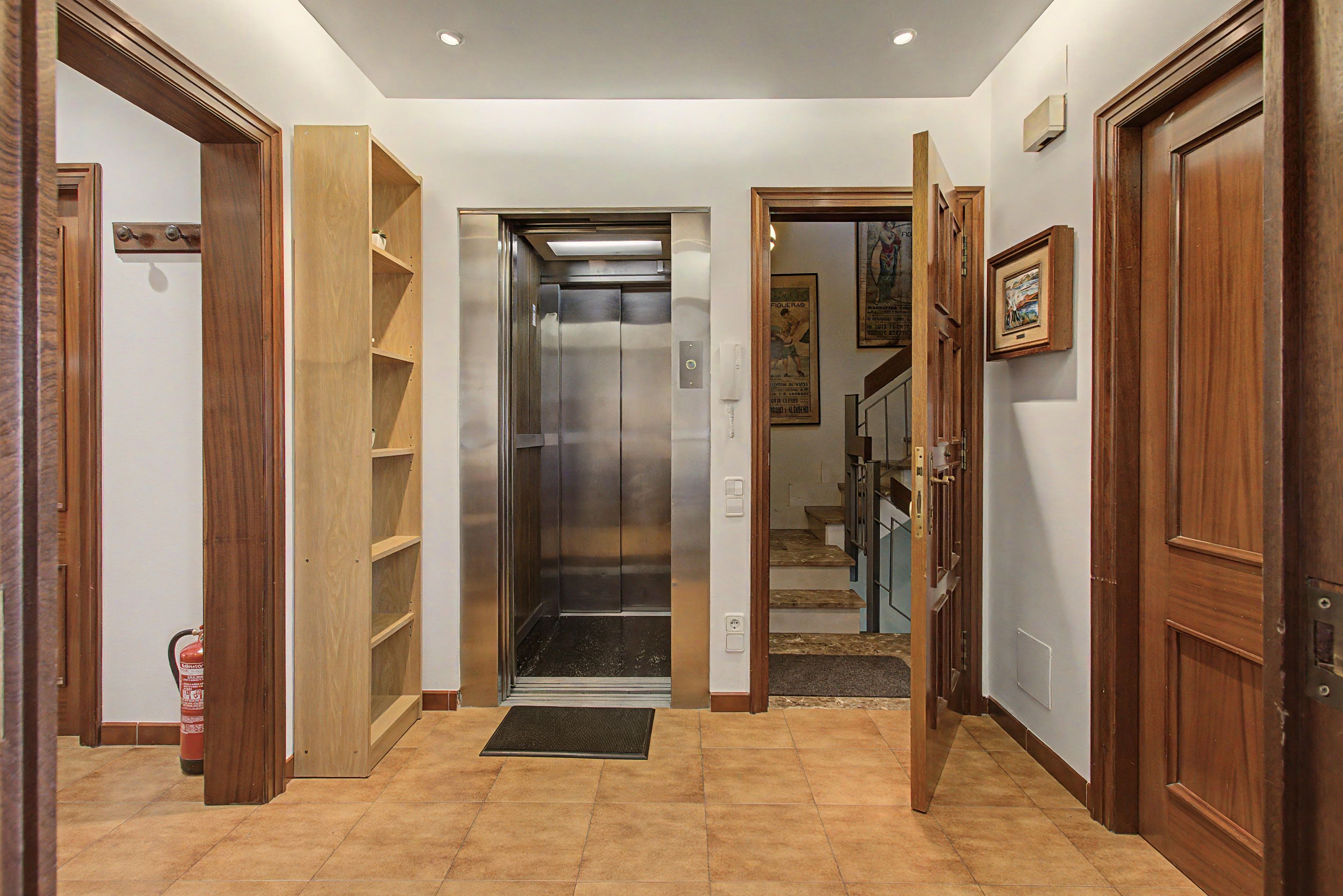 Un ascensor con acabado metálico, flanqueado por puertas y estantes de madera, que conduce a una escalera.