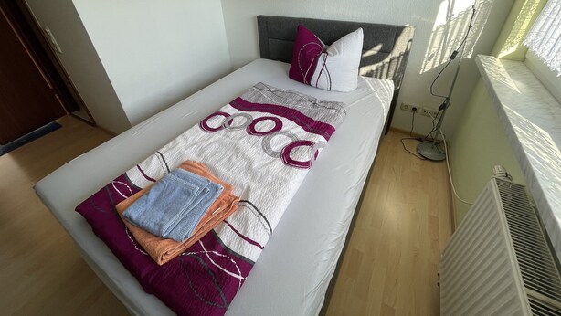 Een netjes opgemaakt bed met een geborduurde deken, kussens en opgevouwen handdoeken.