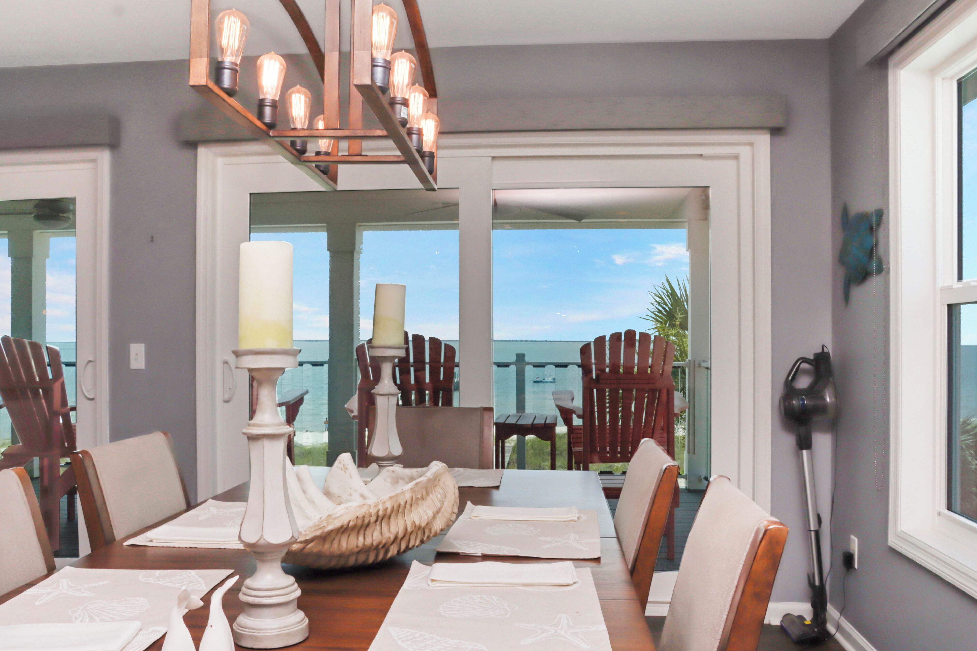 A dining area with a table set for four, a chandelier, and a view of the sea through large windows.