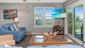 Ein Wohnzimmer mit einem blauen Sofa, einem Fernseher, einem Deckenventilator und einem großen Fenster mit Blick auf den Strand.