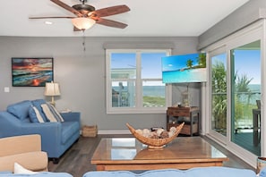Ein Wohnzimmer mit einem blauen Sofa, einem Fernseher, einem Deckenventilator und einem großen Fenster mit Blick auf den Strand.