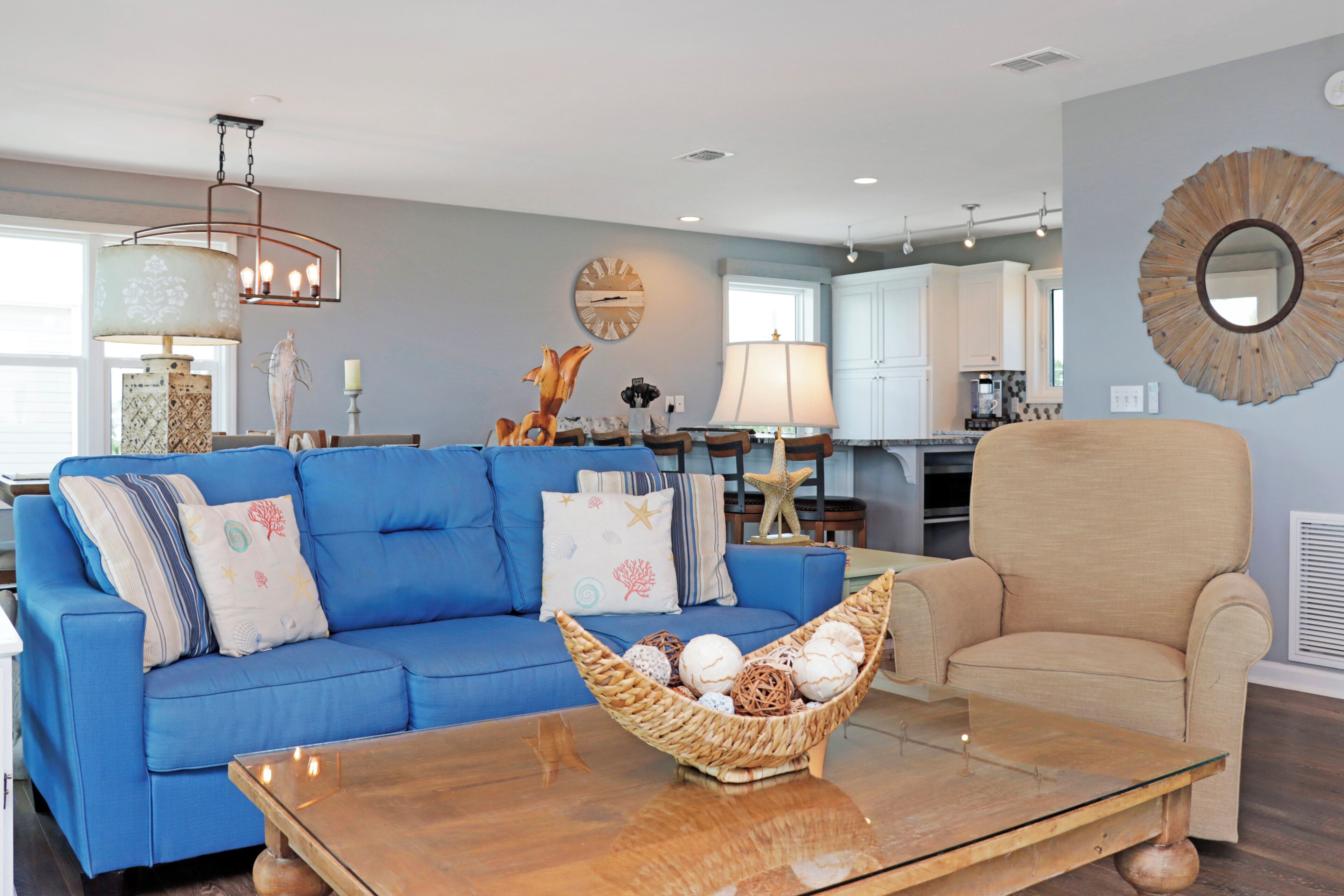 Un salon avec un canapé bleu, un fauteuil beige et une table basse en bois, et une cuisine en arrière-plan.