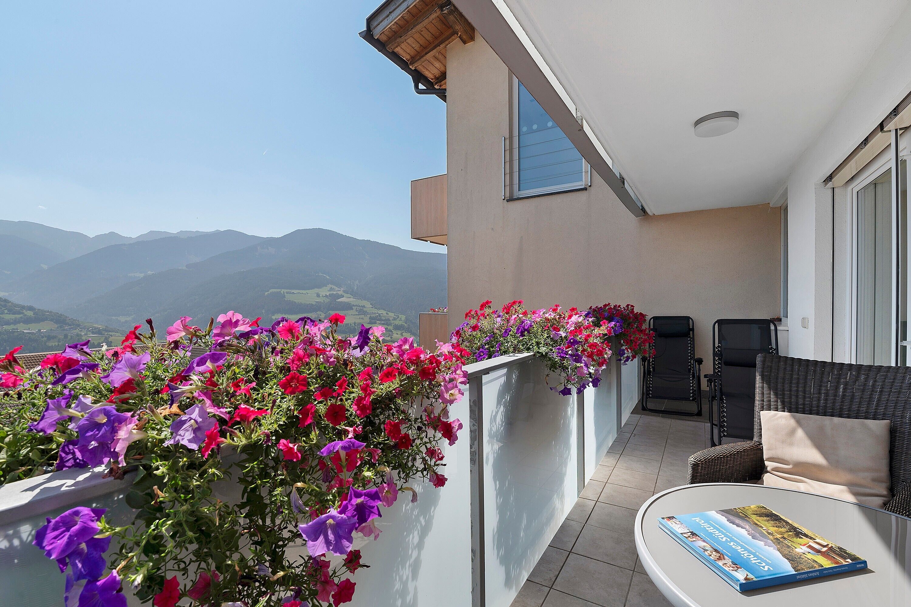 Un balcon avec vue sur les montagnes, une chaise, une table avec un livre et une jardinière remplie de fleurs.