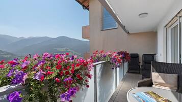 Un balcon avec vue sur les montagnes, une chaise, une table avec un livre et une jardinière remplie de fleurs.
