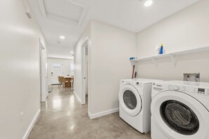 A laundry room with two front-loading washing machines, a dining area with chairs, and a hallway leading to another room.