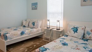 Un dormitorio con dos camas, cada una con una colcha de motivos de tortuga marina, una ventana con persianas y una pequeña mesa con dos lámparas.