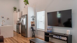 Una sala de estar moderna con televisor de pantalla plana, un banco de madera y una zona de cocina con armarios blancos y un refrigerador.