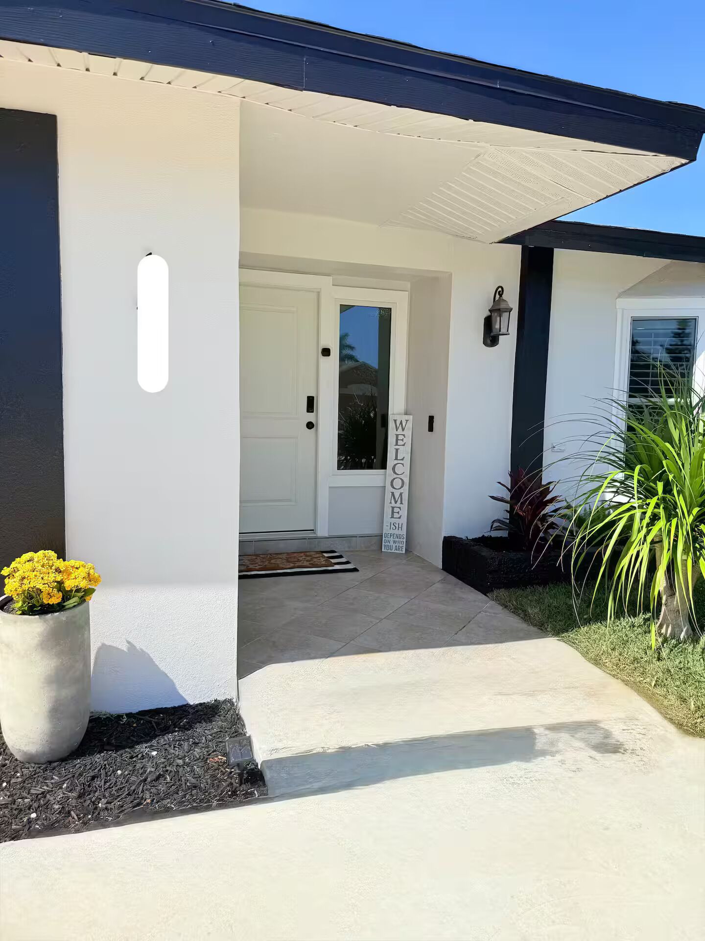 A modern house entrance with a white door, a welcome sign, and a potted plant.