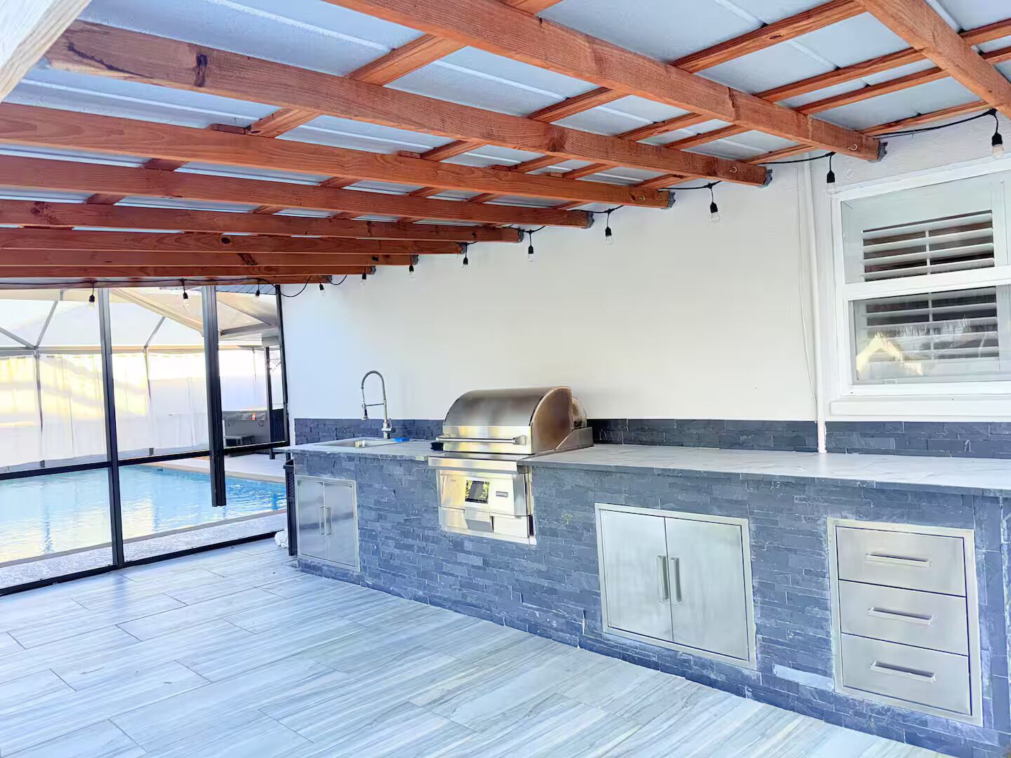 An outdoor kitchen area with a built-in grill, a sink, and a window overlooking a pool.