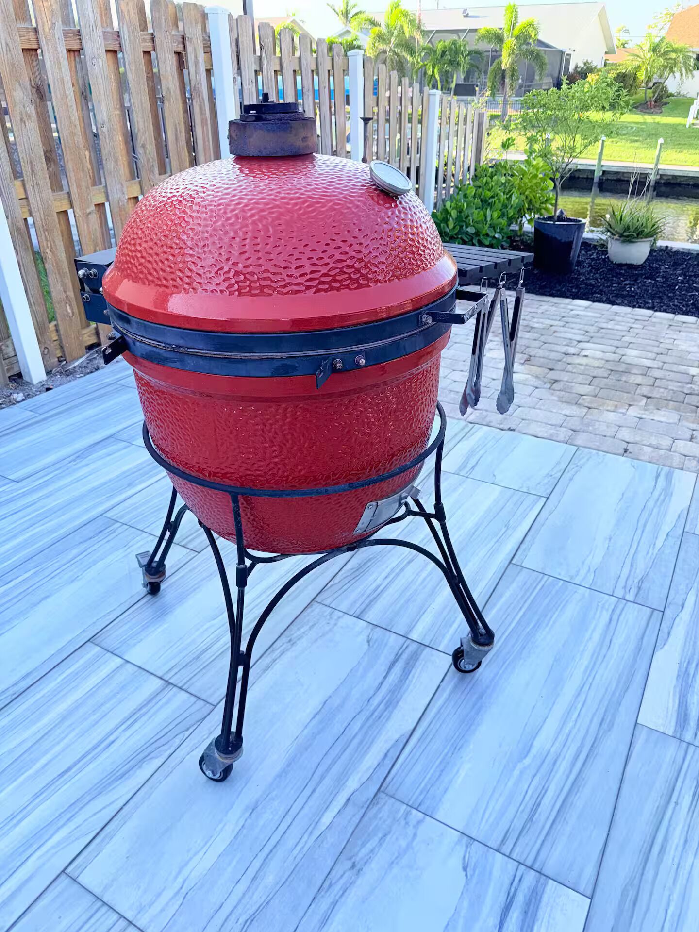 A red barbecue grill on a patio with a wooden fence and plants in the background.