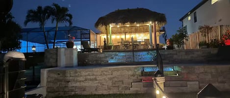 A pool area with a thatched-roof structure, outdoor seating, and a stone wall.