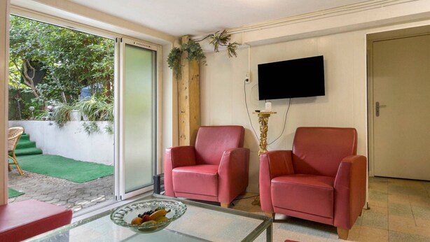 Una habitación con dos sillas rojas, una mesa de vidrio con fruta, un televisor en la pared y un ventanal corredizo que da a un jardín.